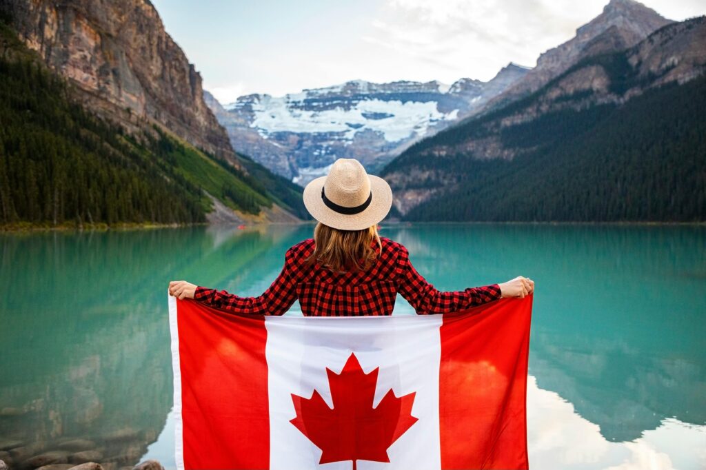 A woman in a red-and-black plaid shirt stands with her back to the camera. She is holding a Canadian flag behind her as she looks at a view of an emerald lake in front of the Rocky Mountains.